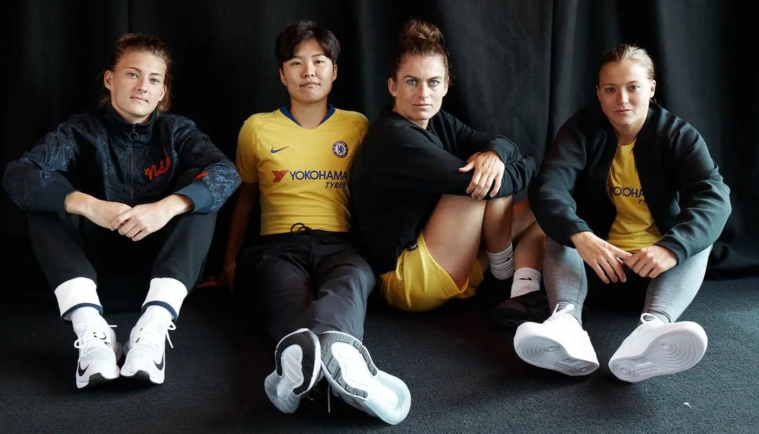 Four Chelsea FC Women players sitting against a black curtain in Nike apparel and the yellow 2018/19 away kit