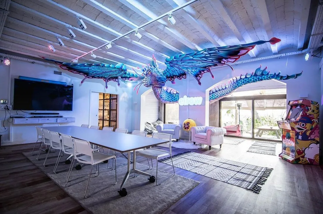 elrow headquarters meeting room: a long table under a giant sculpted dragon, with a neon elrow sign and an arcade machine